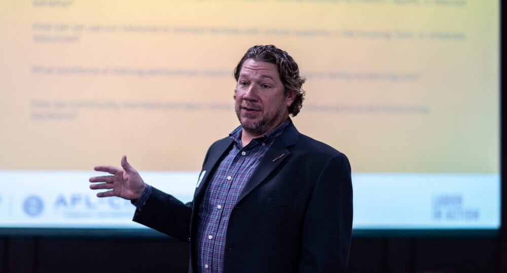 Ryan Harmon speaks in a black blazer at Labor in Action in front of a large presentation screen.