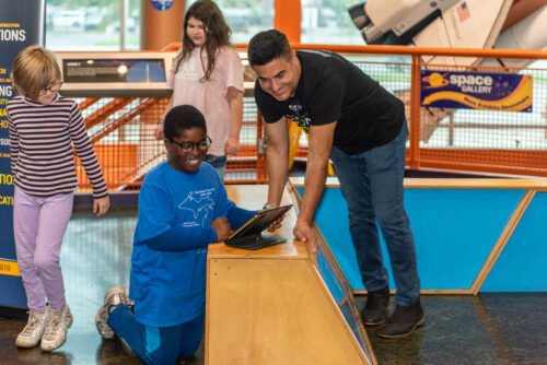 Volunteer helps student control robots from a tablet.