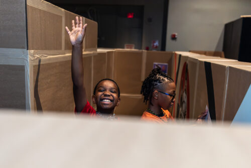 Two students in cardboard maze: one smiles and waves while the other sketches a map.